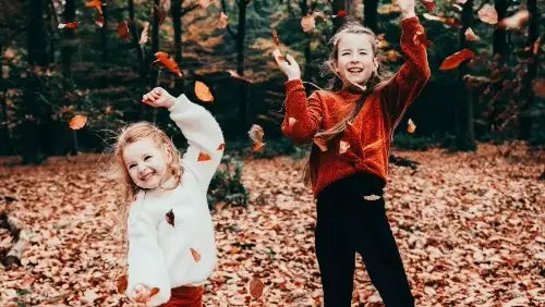 Twee kinderen in herfstkleding spelen speels met bladeren in een bos, de één draagt een oranje trui en de ander een witte trui met bladpatronen.